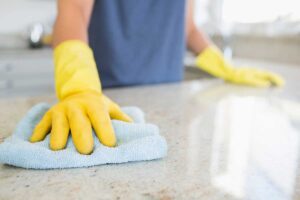 Person dusts furniture while a vacuum and cleaning supplies wait nearby in a bright home.