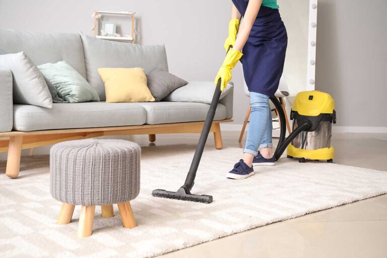 Person using a microfiber cloth to dust furniture while a vacuum and cleaning supplies sit nearby.
